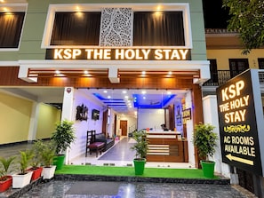 Entrada de un hotel con mostrador de recepción, habitaciones con aire acondicionado disponibles y un cartel que indica el nombre del hotel 'KSP THE HOLY STAY'.