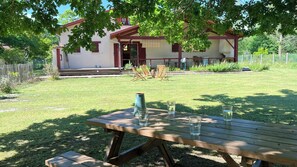 Ein Holztisch für ein Picknick mit Gläsern und einer Vase, ein Rasen und ein Gebäude mit roter Tür und Fenstern.