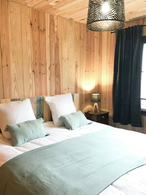 Ein Zimmer mit Holzwandverkleidung, einem Bett, Kissen, einem Nachttisch, einer Lampe und einem Fenster mit Vorhängen.