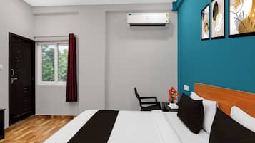 A hotel room with a bed, a desk with a chair, an air conditioning unit, a window with curtains, and artwork on the wall.