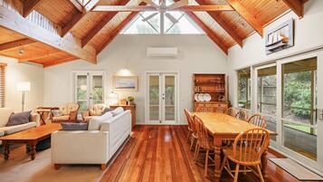A spacious living area with a high ceiling, wooden beams, a dining table, and a view through large windows.