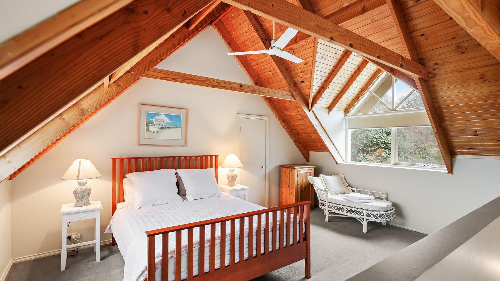 A bedroom with a sloped ceiling, a bed, bedside lamps, a chair, and a window.