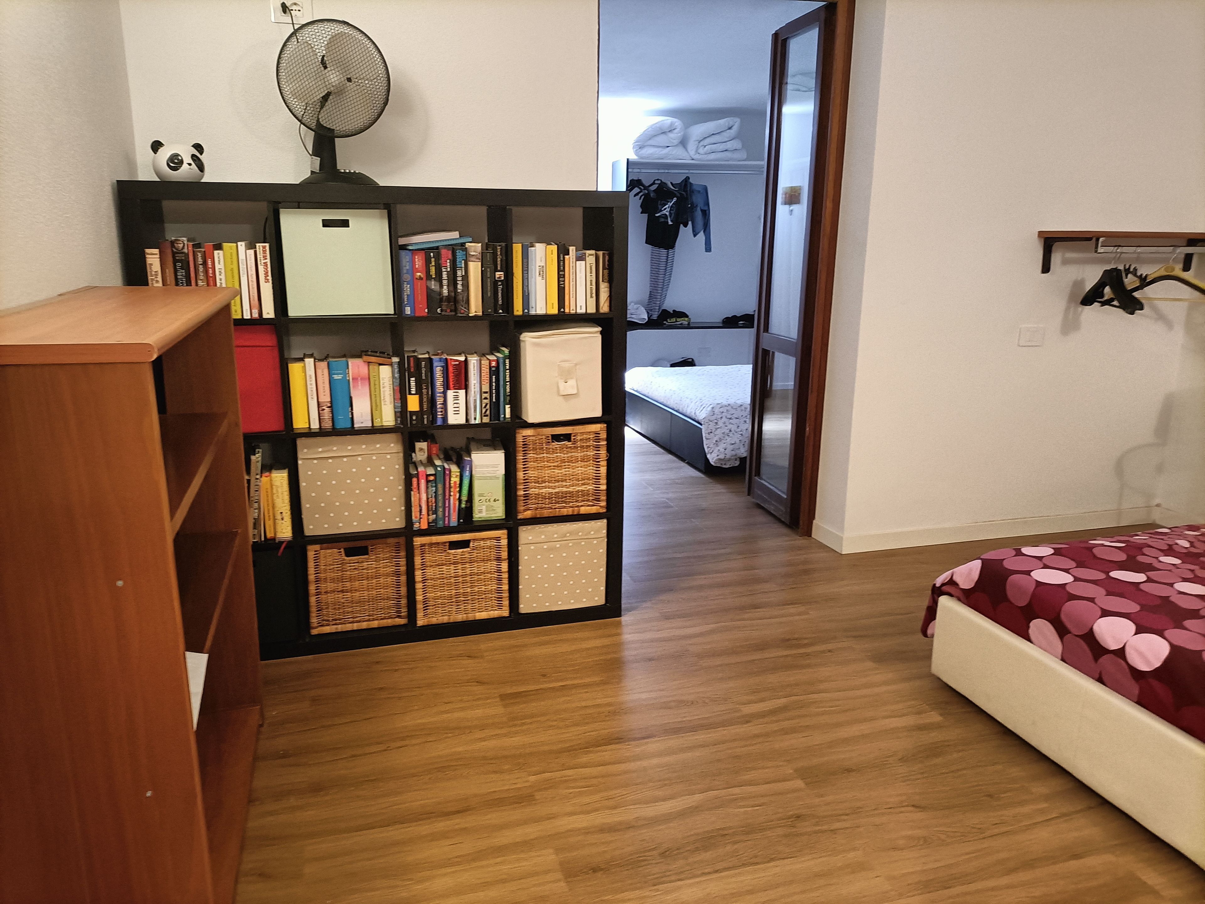 Una habitación con una estantería de madera, una cama con colcha de lunares y un armario con ropa colgada.