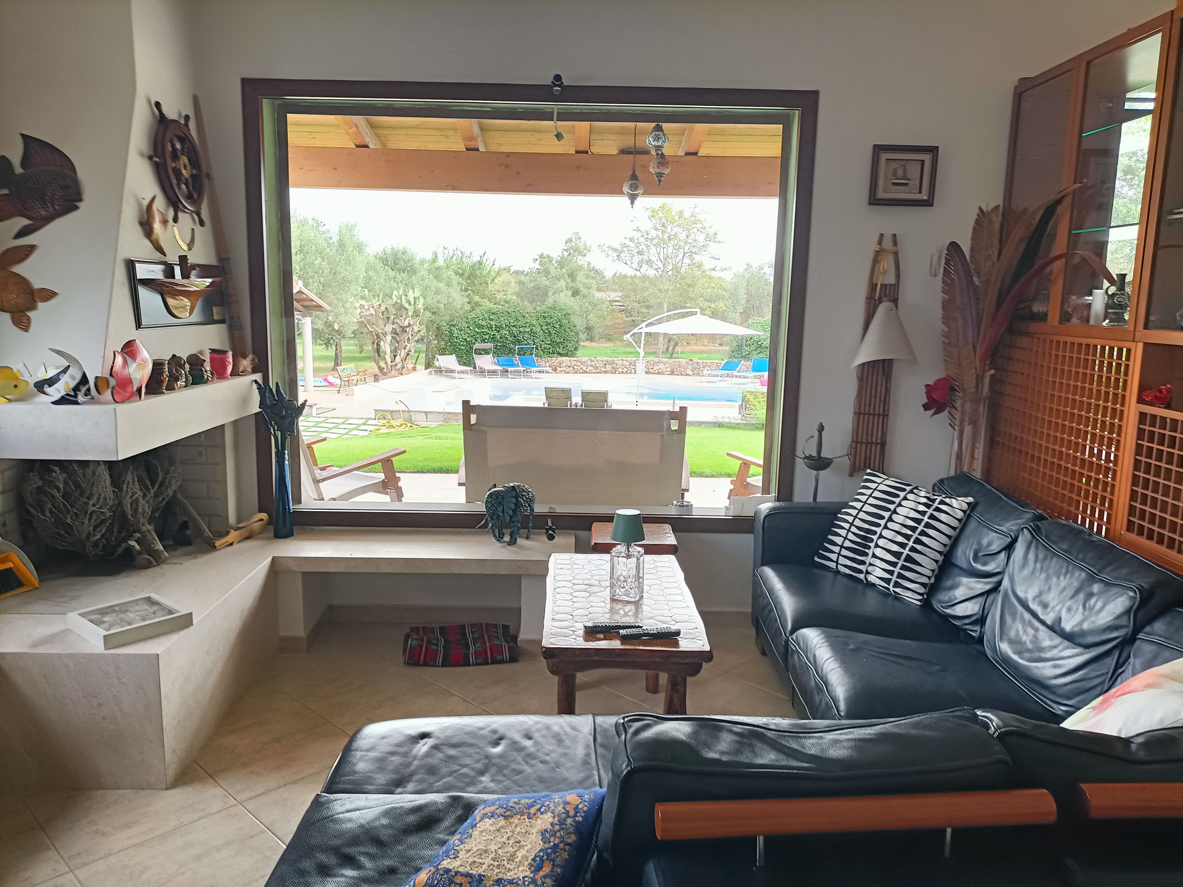 Sala de estar con vista a una piscina y jardín, un sofá de cuero negro, una mesa de centro y una estantería con objetos decorativos.