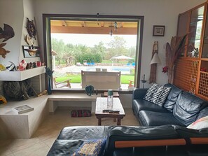 Sala de estar con vista a una piscina y jardín, un sofá de cuero negro, una mesa de centro y una estantería con objetos decorativos.