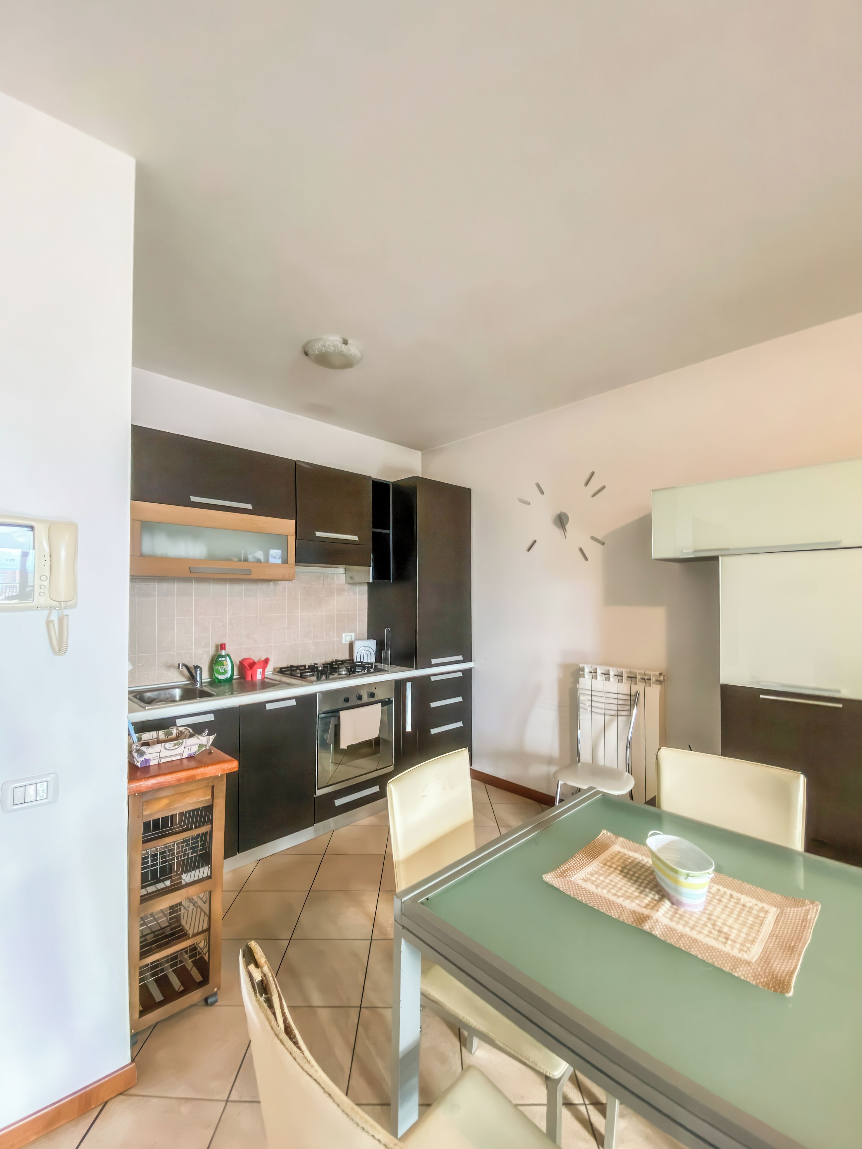 A modern kitchen with a dining area, including a table with chairs, a clock on the wall, and a built-in oven.