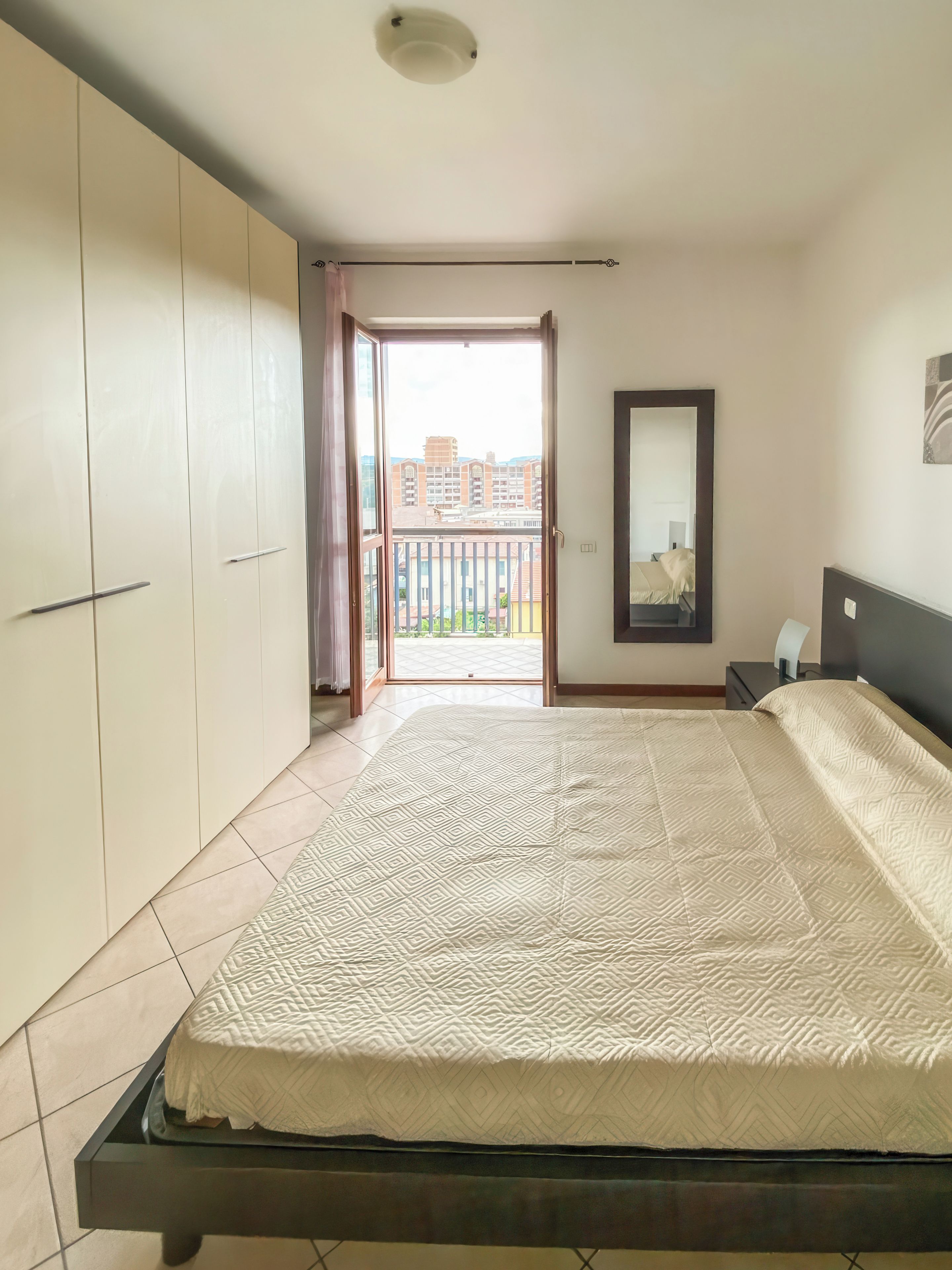Une chambre à coucher avec un lit, une armoire et un balcon donnant sur des immeubles.