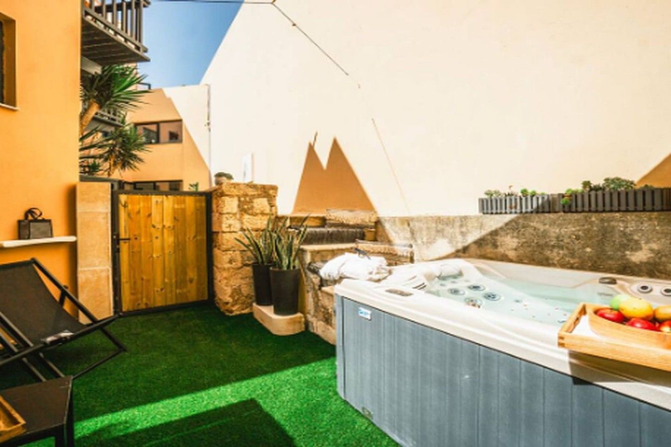 A hot tub in a backyard with a stone wall, a wooden gate, and a small table with fruit.
