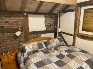 Ein Schlafzimmer mit einem hölzernen Kopfende, einer Ziegelwand, einer Holzbalkendecke, einem Bett mit einer karierten Steppdecke, einem Nachttisch mit einer Lampe und einem Fenster mit Holzjalousien.