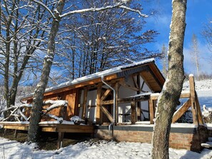 Eine Holzhütte mit geneigtem Dach, umgeben von schneebedeckten Bäumen und blauem Himmel.