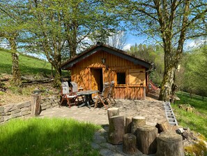 Una cabaña de madera con un patio de piedra, mobiliario exterior y un entorno rústico rodeado de árboles.