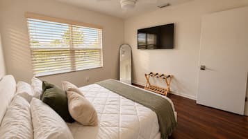 A bedroom with a bed, a TV mounted on the wall, a window with blinds, and a door.