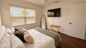 Ein Schlafzimmer mit einem Bett, einem an der Wand befestigten Fernseher, einem Fenster mit Jalousien und einer Tür.