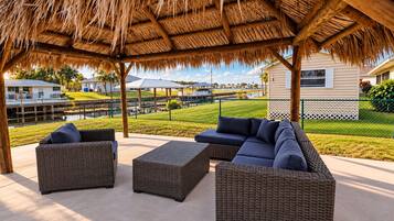 An outdoor patio with wicker furniture, a thatched roof, and a view of a residential area with houses and a body of water.