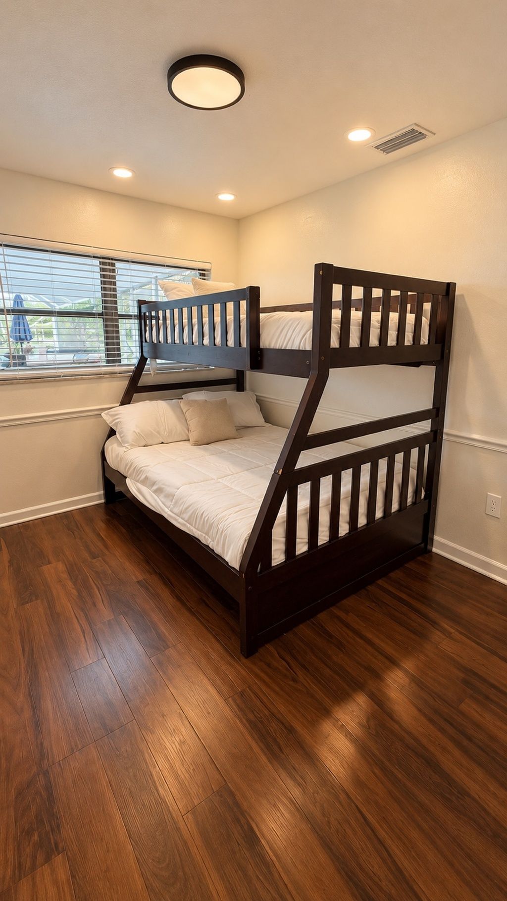 A bunk bed with a wooden frame and white bedding in a room with wooden flooring and a window with blinds.