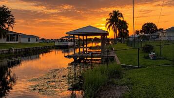 A wooden dock extending into a calm body of water with lily pads, a gazebo, and a sunset in the background.