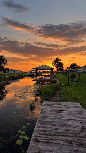 A wooden dock extending into a calm body of water with lily pads, a gazebo, and a sunset in the background.