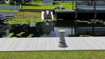 Un muelle de madera que lleva a un estanque tranquilo con lirios acuáticos, un pedestal de piedra y un bote cubierto.
