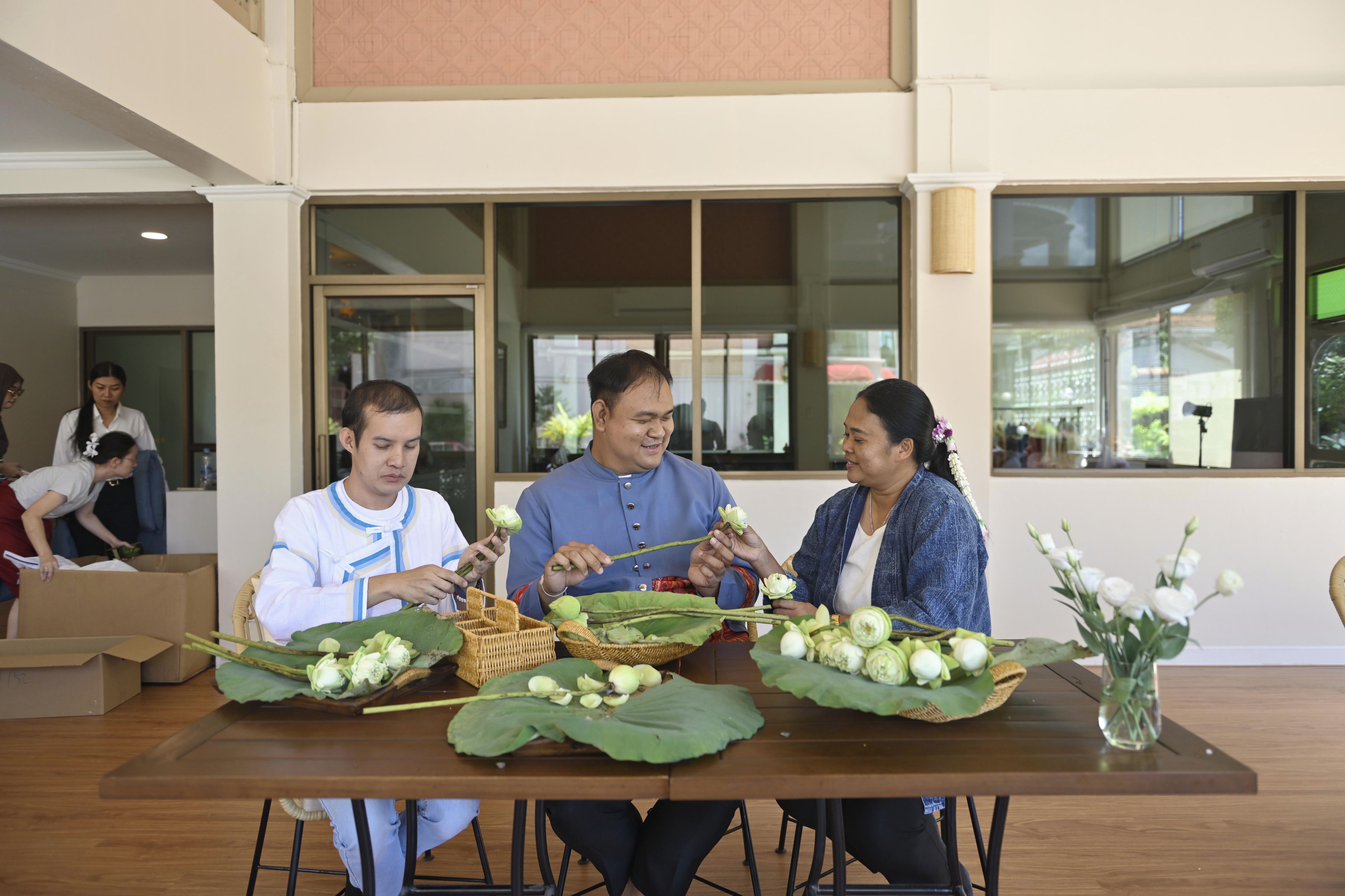 A group of people seated at a table with large leaves and flowers, possibly engaged in a cultural activity.