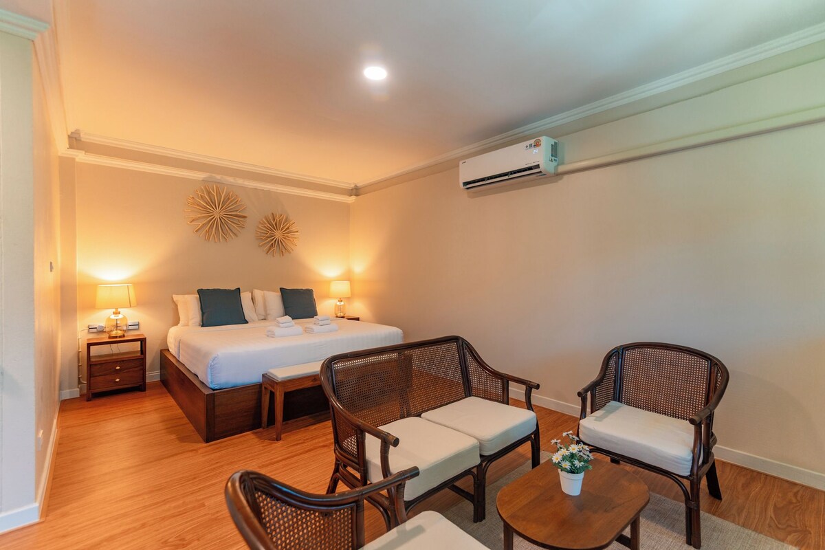 Quarto de hotel com uma cama, duas mesinhas de cabeceira, um abajur, um ar condicionado fixado na parede e duas cadeiras de vime com almofadas.
