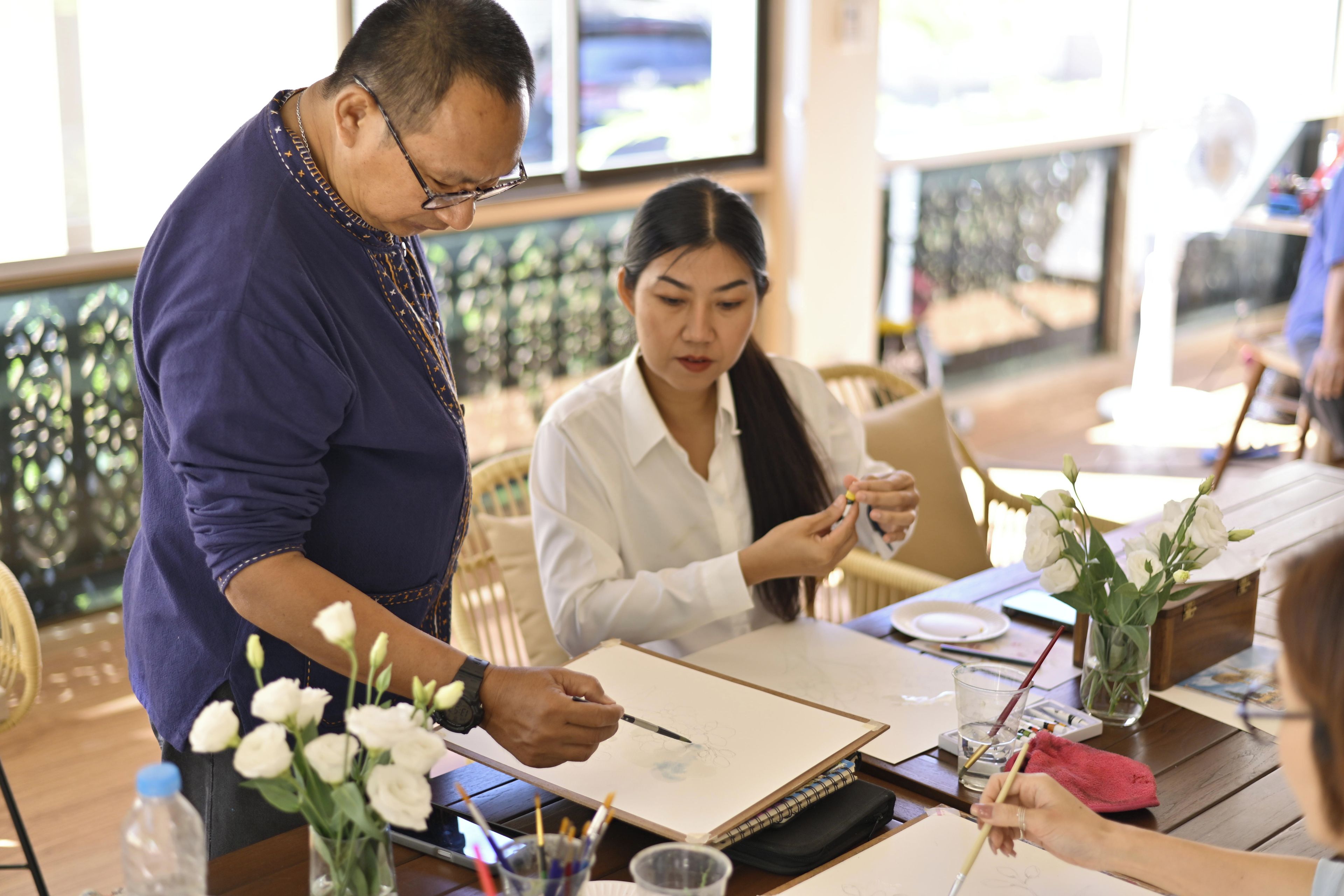 A person drawing on a paper with a pencil, a vase with flowers, and a table with a laptop and a cup.