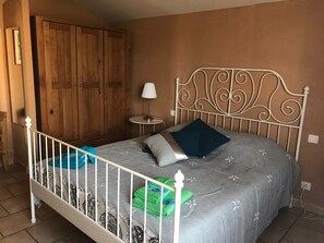A bedroom with a metal-framed bed, a wooden wardrobe, a small round table with a lamp, and a wall-mounted light switch. - House 'La Bastide de l'Olivier' with Provençal Charm and Private Pool at the Foot of Ventoux (Flassan)