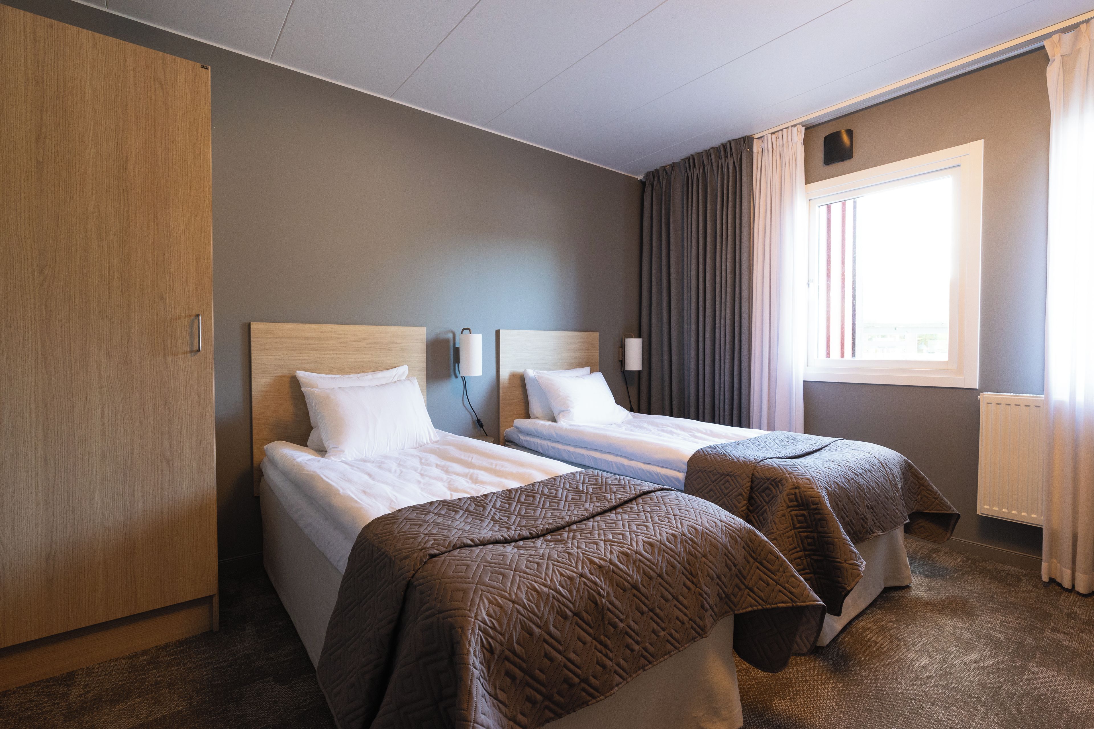 Ein Hotelzimmer mit zwei Betten, einem Kleiderschrank, einem fenster mit Vorhängen und einer Wandlampe.