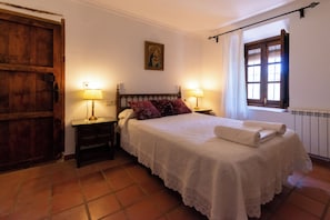 A bedroom with a large bed, two bedside tables, a wooden door, a window with shutters, and a wall-mounted painting.