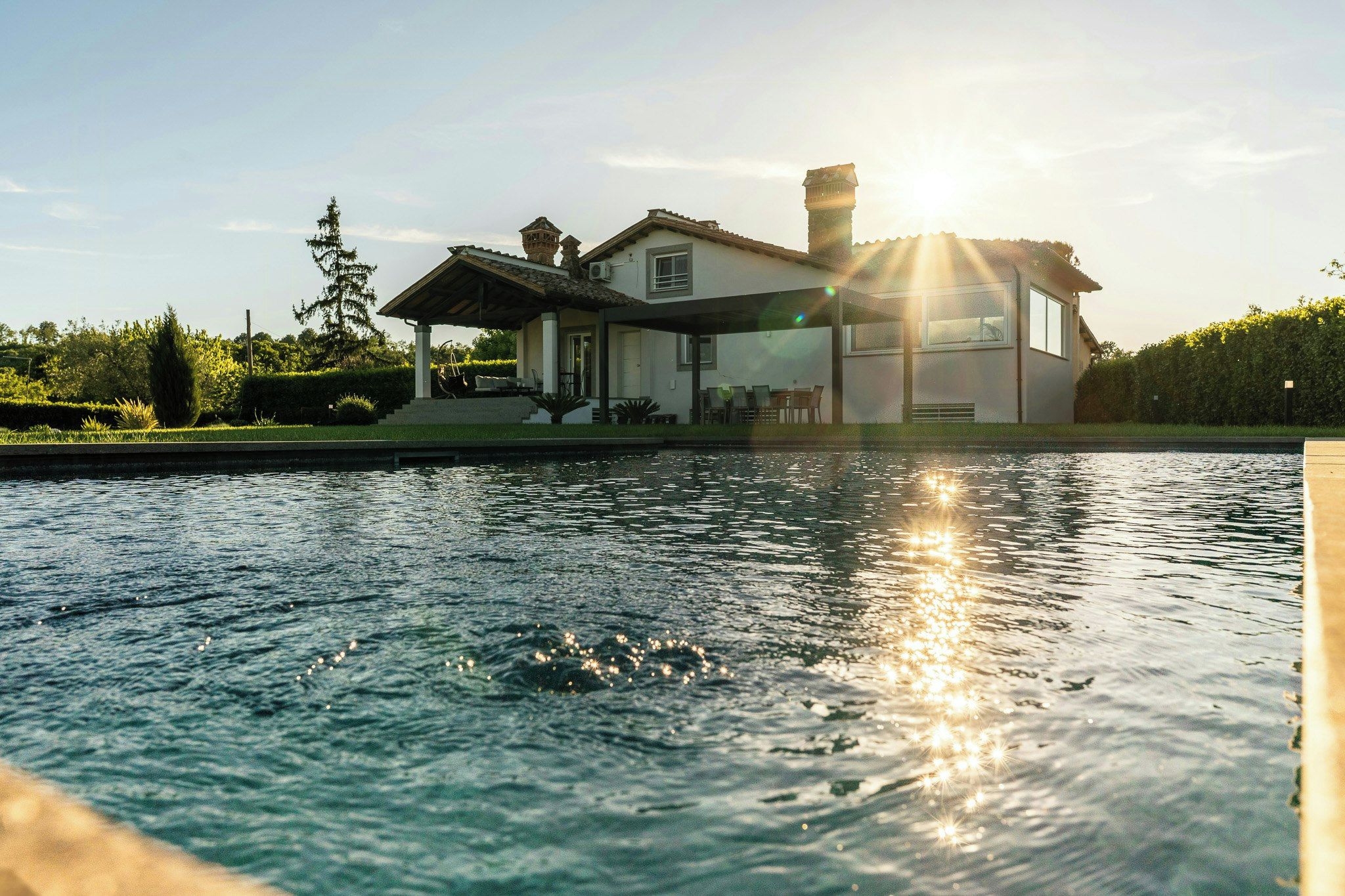 A house with a swimming pool in the foreground and a sun setting in the background.