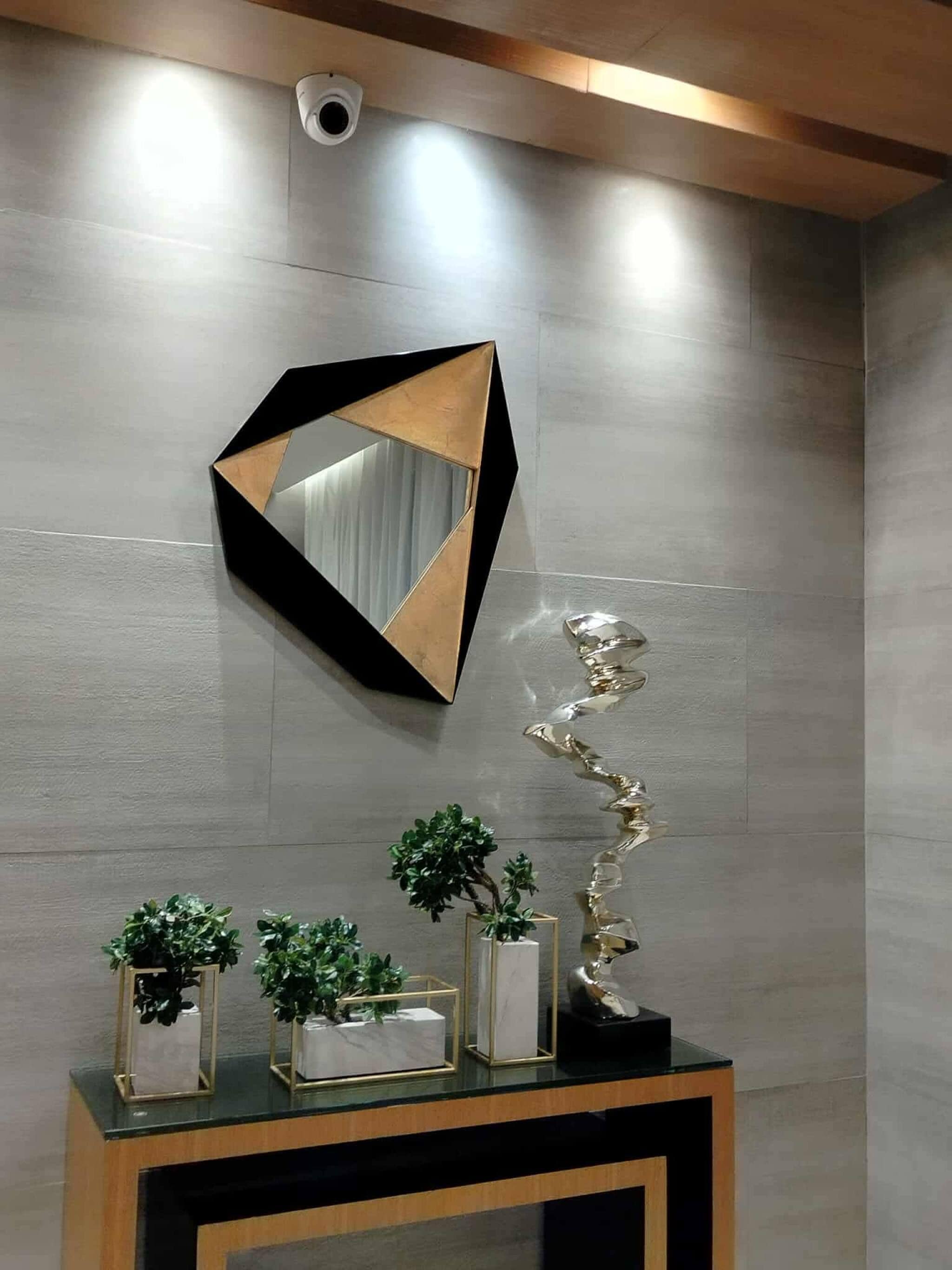 Un interior moderno con un espejo geométrico, una escultura metálica y plantas en macetas sobre una consola.