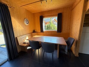 Ein Interieur einer Holzhütte mit einem Esstisch, zwei Stühlen, einer Uhr und einem Fenster mit Vorhängen.