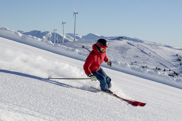 En person står på ski ned ad en sneklædt skråning med klar himmel og bjerge i baggrunden.