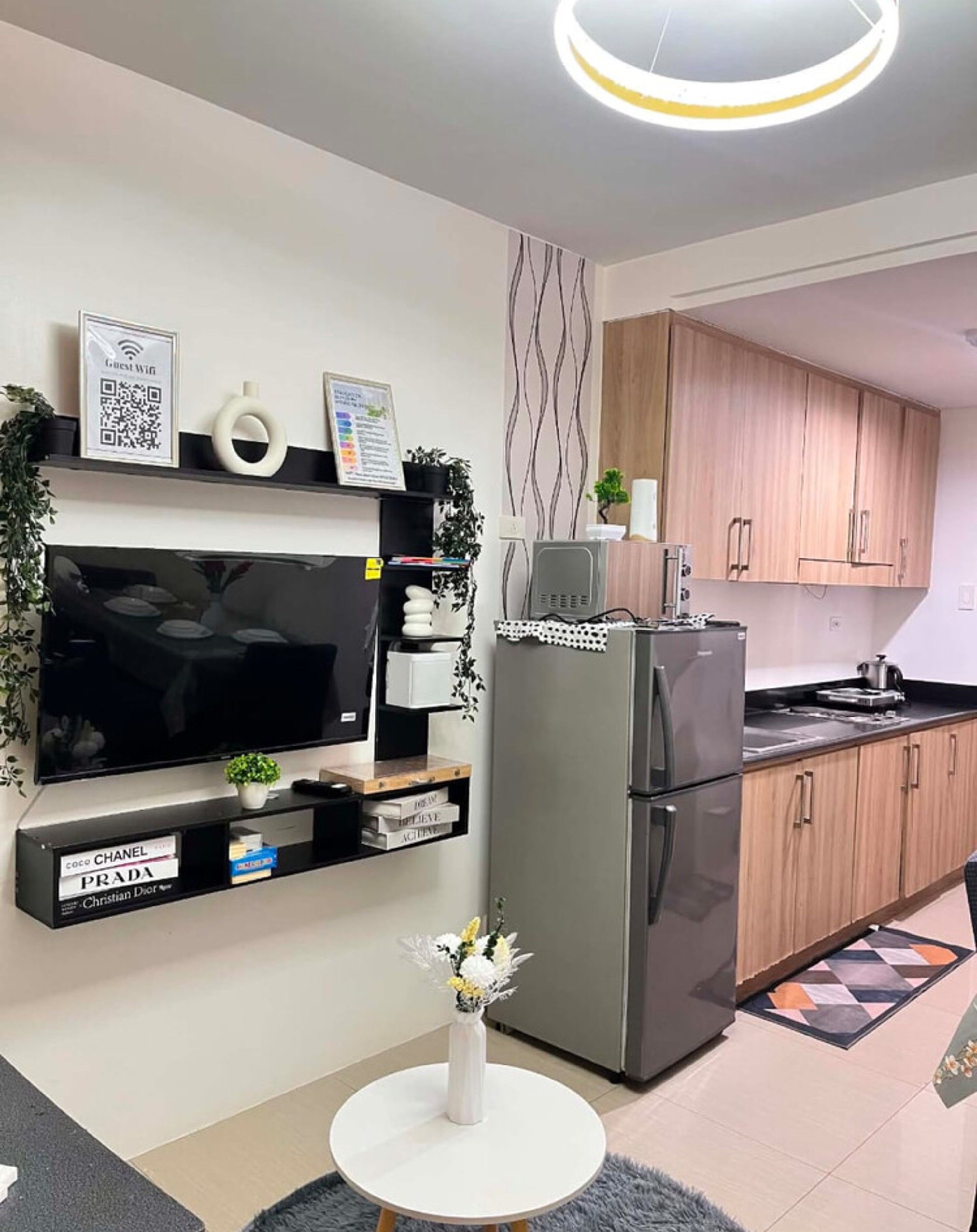 A modern kitchen with a flat-screen TV, a refrigerator, wooden cabinets, and a small dining table with a vase of flowers.
