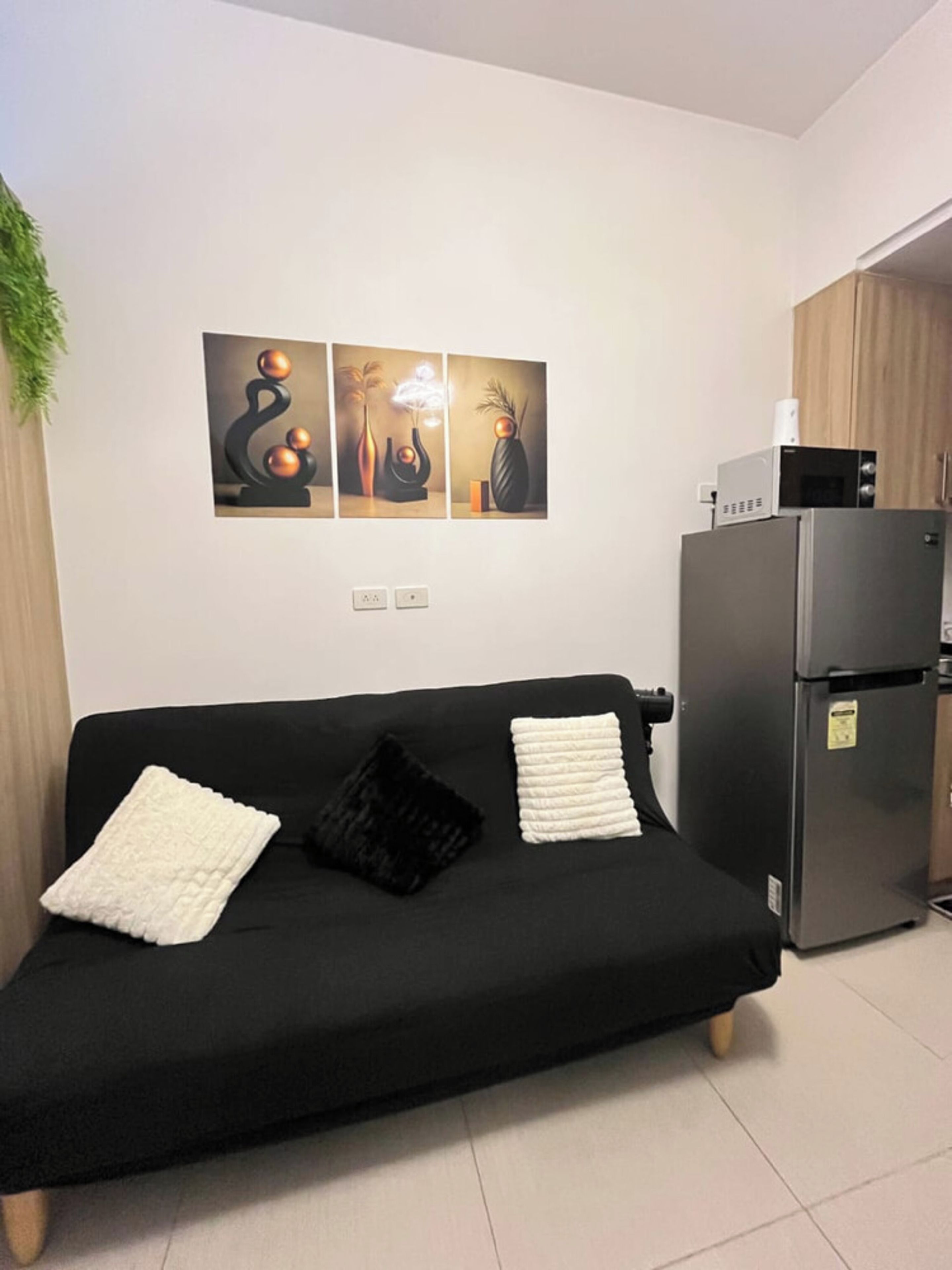 A modern living room with a black sofa, two cushions, a wall-mounted artwork, a kitchen area with a refrigerator, and a microwave.