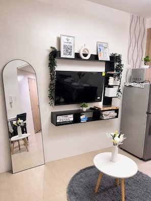 Una sala de estar moderna con un televisor de pantalla plana, un espejo grande, una mesita redonda con un florero y un refrigerador.