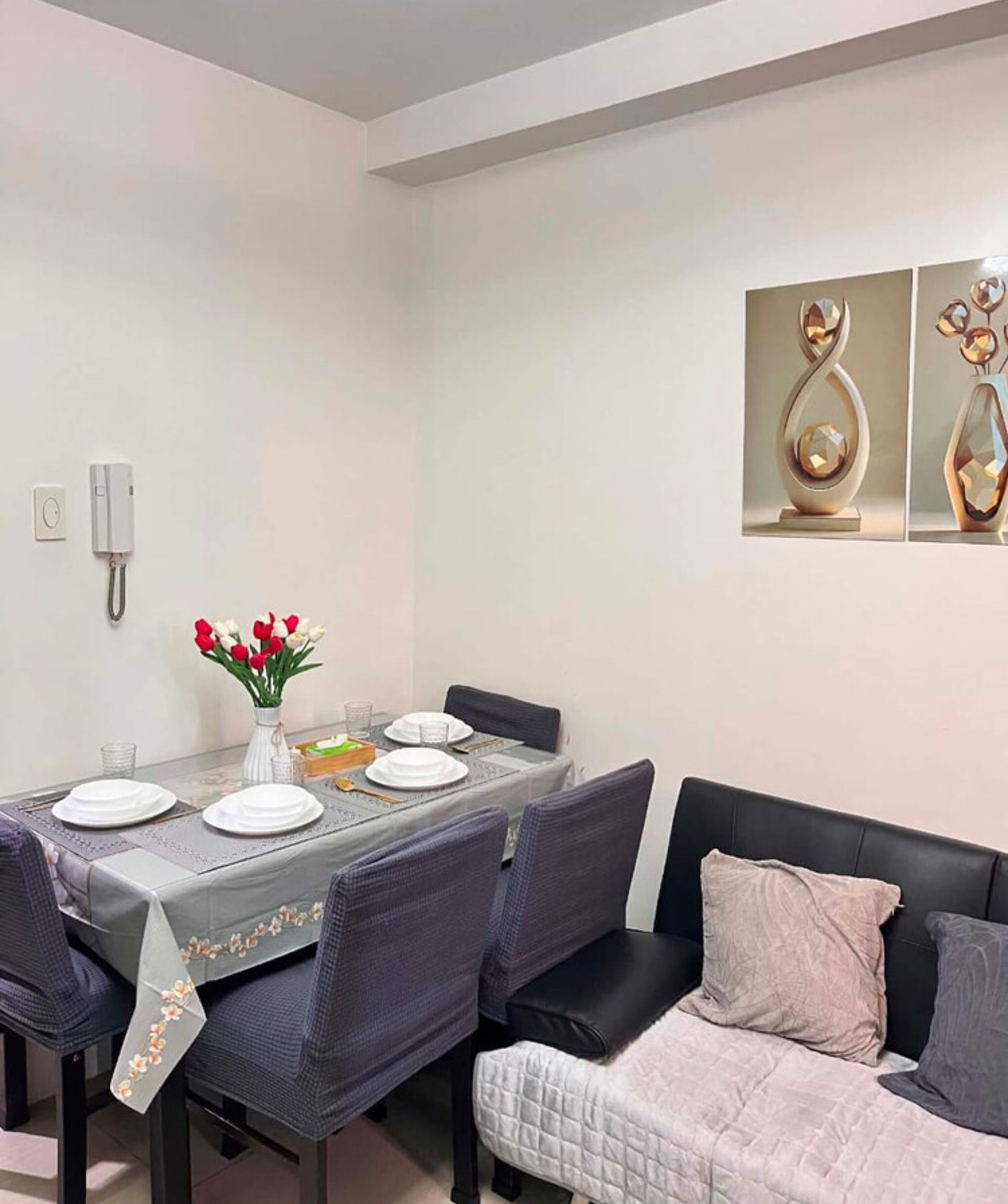 A dining area with a table set for four, chairs, a vase with flowers, and a sofa with cushions.