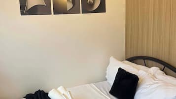 Habitación de hotel con una cama, almohadas, una mesita de noche con un dispensador de pañuelos de papel y un objeto verde, y una pared con una gran letra metálica 'S'.