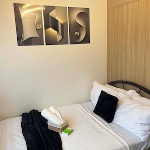 Habitación de hotel con una cama, almohadas, una mesita de noche con un dispensador de pañuelos de papel y un objeto verde, y una pared con una gran letra metálica 'S'.