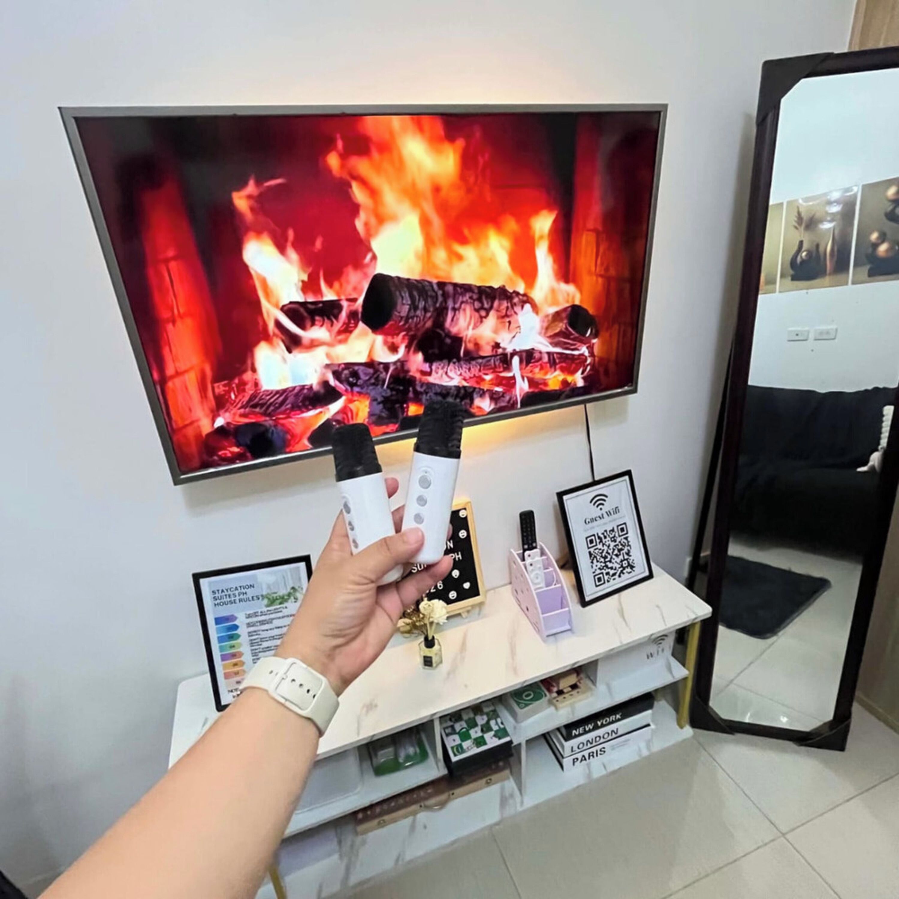 A hand holding two remote controls in front of a television displaying a fire. Below the TV, there is a shelf with various items including a framed QR code, a small vase, and a box labeled 'NEW YORK LONDON PARIS'.