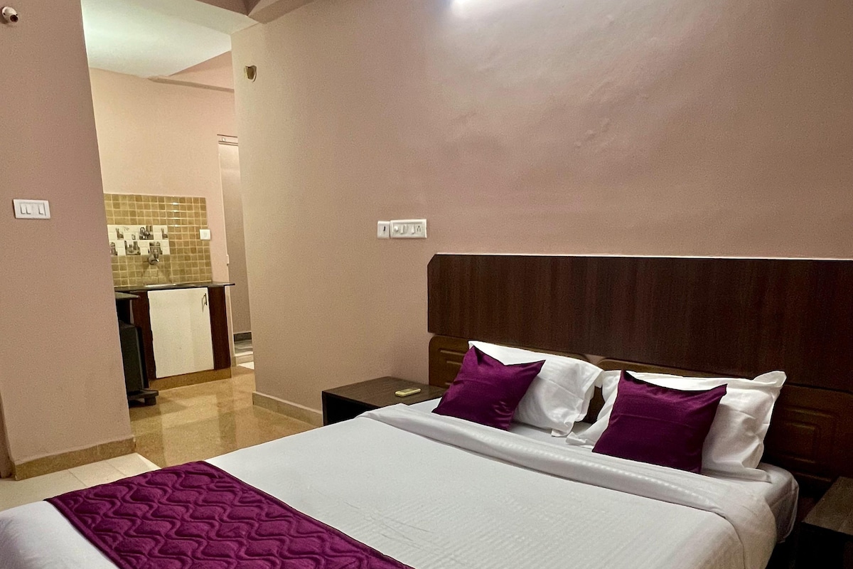 A hotel room with a large bed, a nightstand, a wooden headboard, and a view of the kitchen area.
