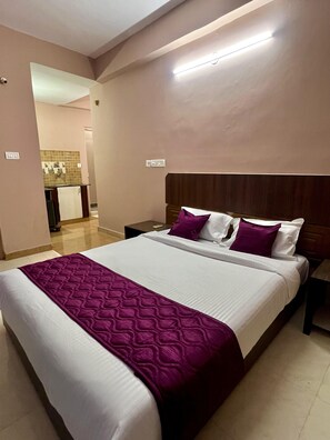 A hotel room with a large bed, a nightstand, a wooden headboard, and a view of the kitchen area. - Namastay signature (Bangalore)