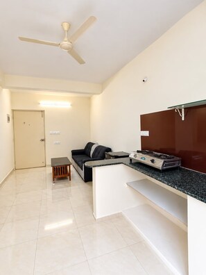 Una habitación con ventilador de techo, un sofá, una mesita y una kitchenette con estufa, además de una puerta.
