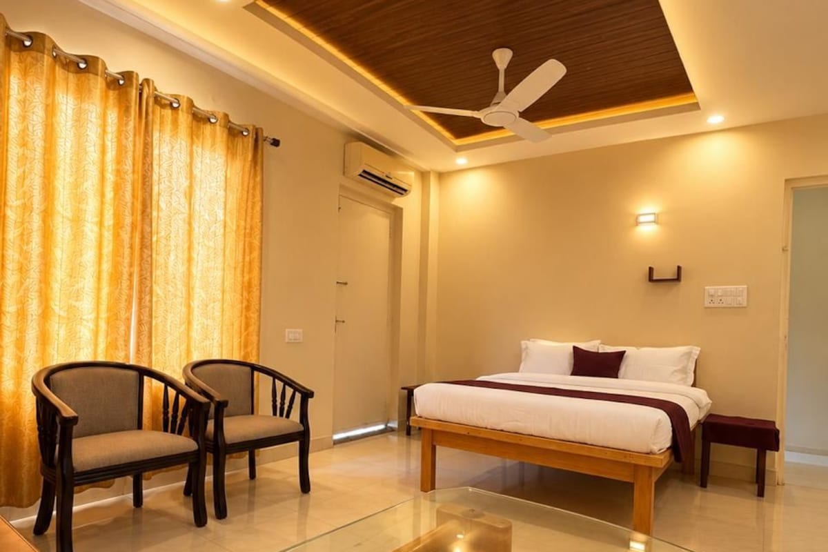 A modern hotel room with a bed, two chairs, a glass coffee table, a ceiling fan, and a wooden ceiling panel.