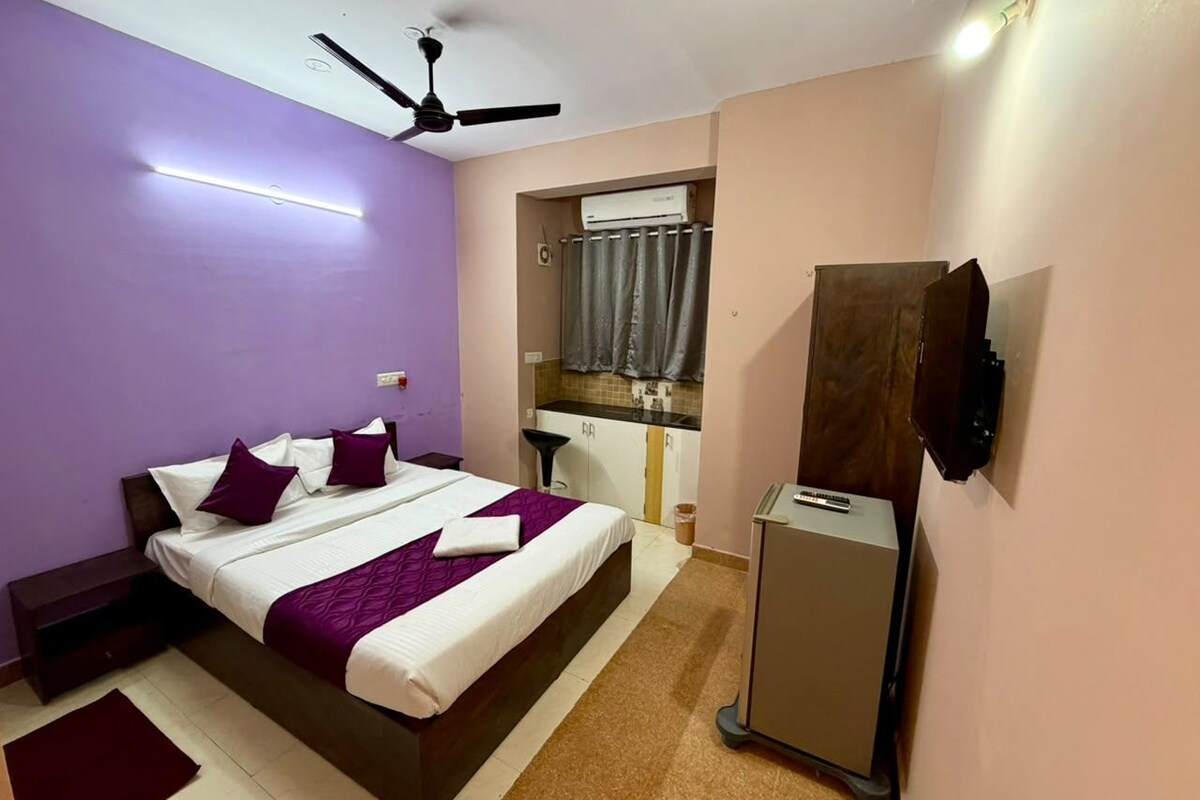 A bedroom with a bed, a ceiling fan, an air conditioning unit, a small kitchenette, and a TV mounted on the wall.