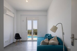 A modern living room with a sofa, a chair, a lamp, and a view of a swimming pool through glass doors. - Villa Elies be a part of Crete (Mesi)
