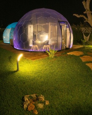 A geodesic dome structure with a glass door, illuminated interior, and a small outdoor area with grass and rocks.