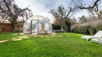 Una estructura con cúpula transparente en un patio trasero, con césped, cerca de madera y dos sillones blancos.