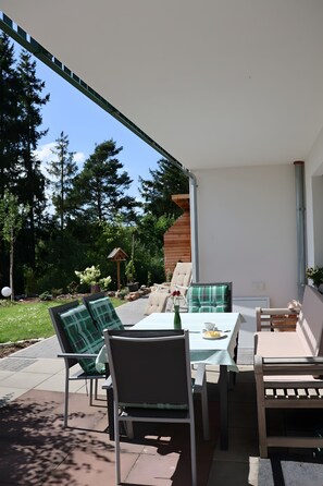 Une terrasse extérieure avec une table et des chaises, entourée de verdure et d’arbres.