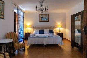 A hotel room with a bed, two bedside lamps, a chair, a table, a chandelier, and a painting on the wall.