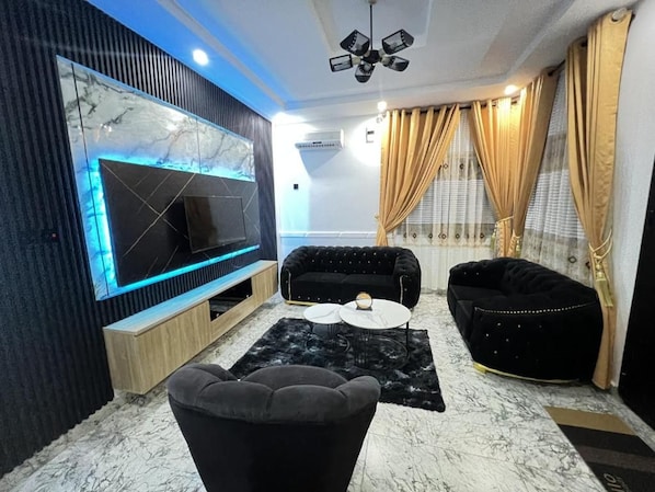 A modern living room with a flat-screen TV, black sofas, a coffee table, and a marble-patterned floor.